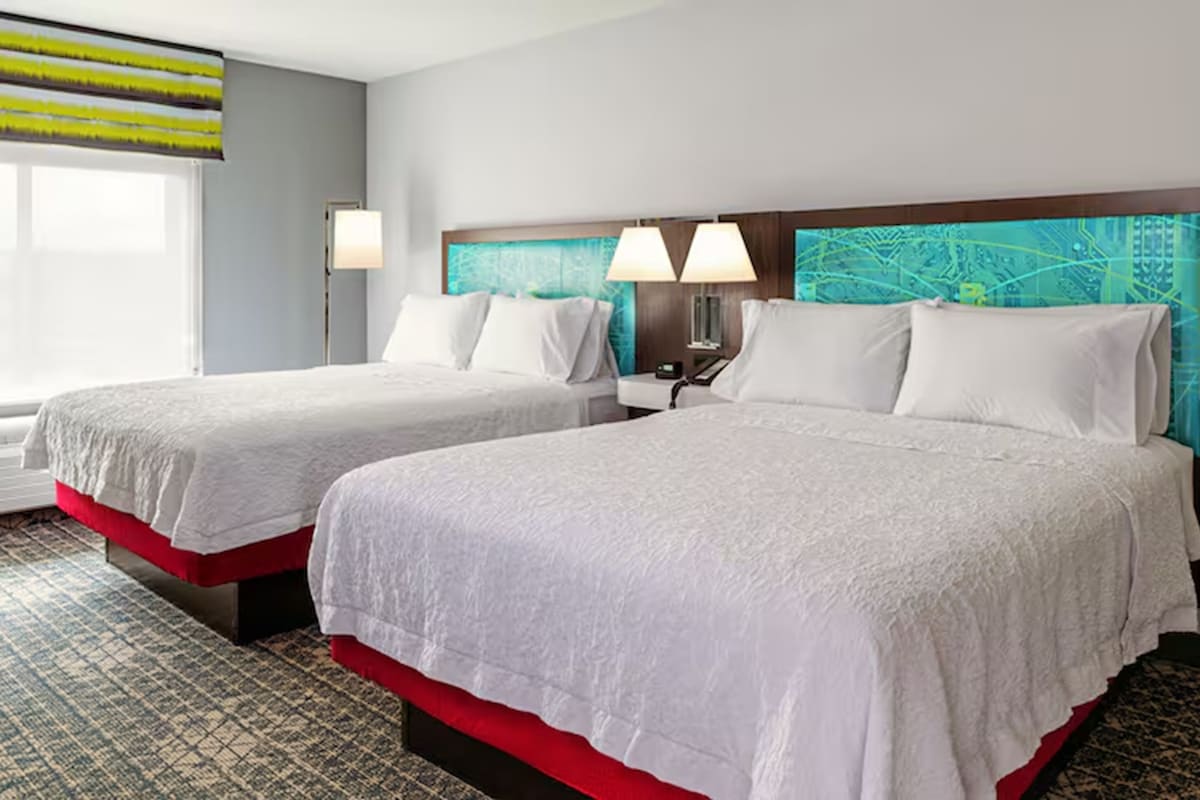 Habitación de hotel con dos camas, un ventanal con persianas, cabecero con diseño de paisaje urbano y alfombra estampada.