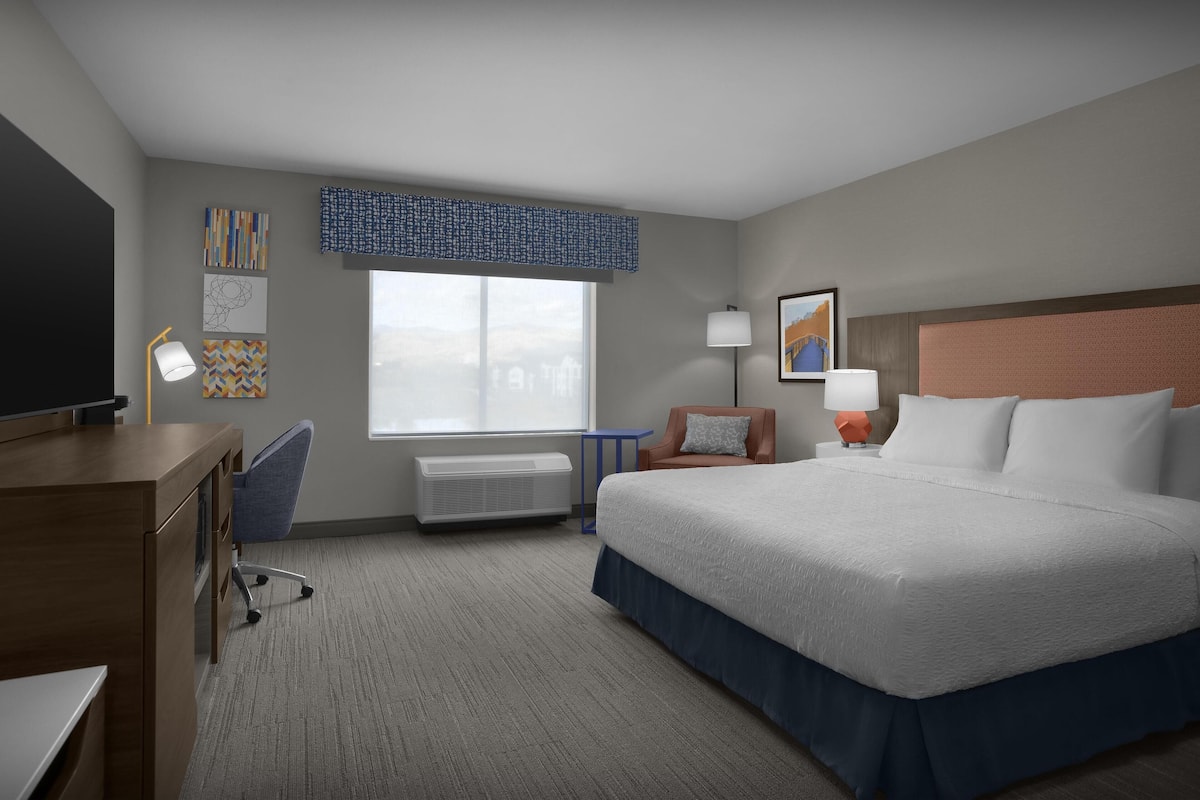 A hotel room with a large bed, a desk with a chair, a window with a view, and artwork on the walls.