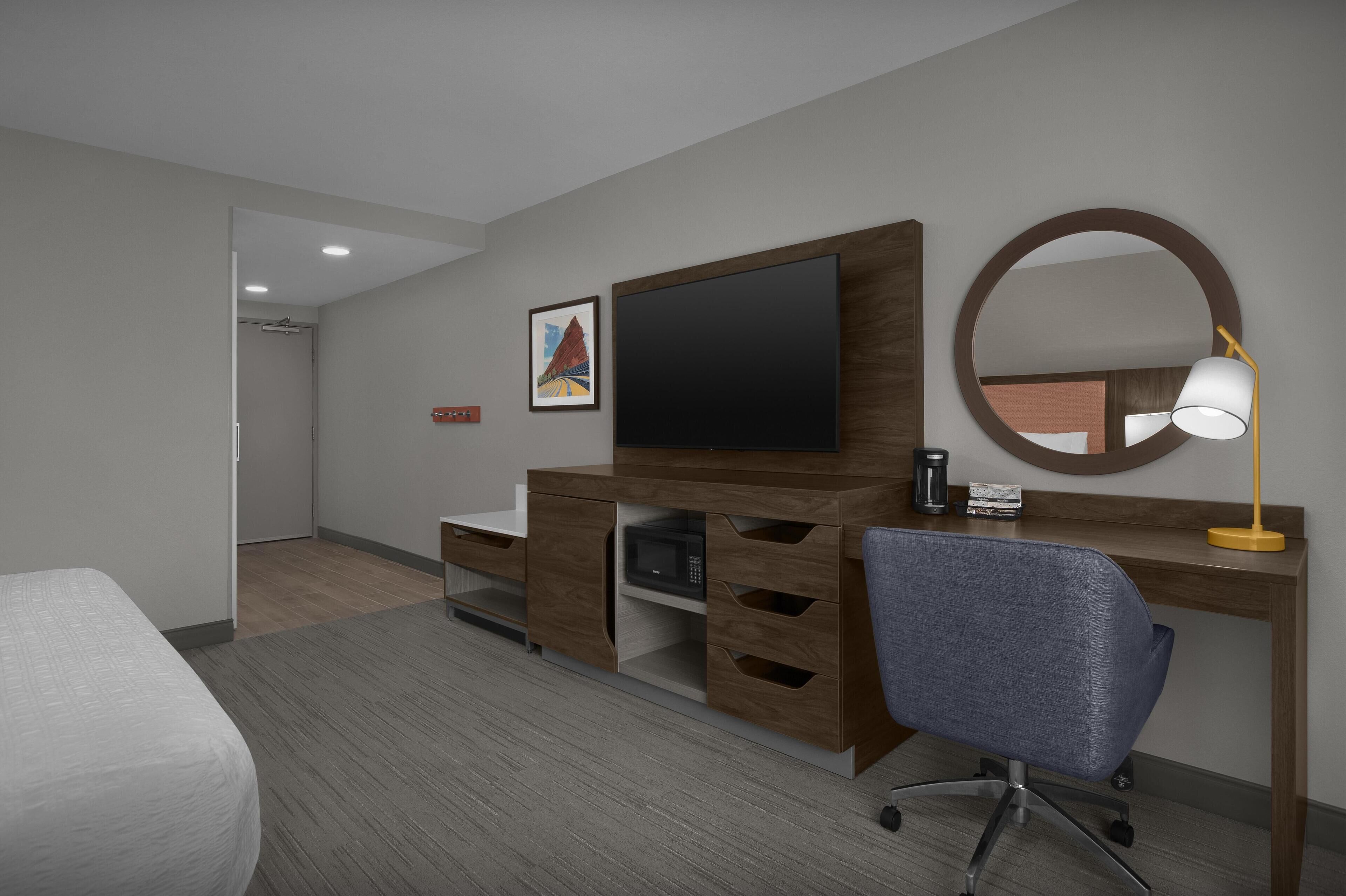 A hotel room with a bed, a desk with a computer, a TV, a microwave, a lamp, and a mirror.