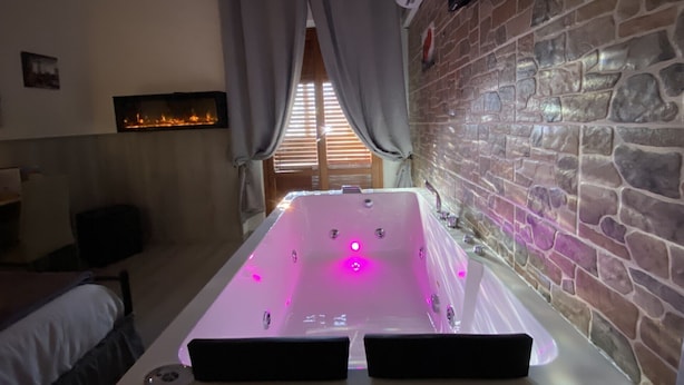 Une salle de bain moderne avec un mur en pierre, une baignoire éclairée de lumière rose et une fenêtre avec des stores.