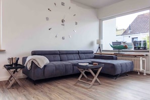 Un salon moderne avec un canapé d’angle gris, une horloge murale, un radiateur et une fenêtre donnant sur une maison.