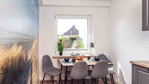 A dining area with a table, chairs, a window, and a view of a house. - Apartment 'Büsumer Butze' with Private Garden and Wi-Fi (Büsumer Deichhausen)