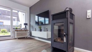 A modern living room with a fireplace, a mounted TV, and a white built-in bench with decorative items. - Apartment 'Büsumer Butze' with Private Garden and Wi-Fi (Büsumer Deichhausen)