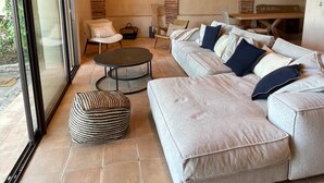 Una sala de estar moderna con un sofá modular grande, una mesa de centro redonda y un reposapiés a rayas. Hay una puerta de cristal que da al exterior, y la habitación cuenta con vigas de madera a la vista en el techo.
