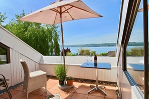 A balcony with a table, chair, and umbrella, overlooking a body of water and greenery. - Holiday Apartment with Lake View, Balcony, and Wi-Fi (Überlingen)
