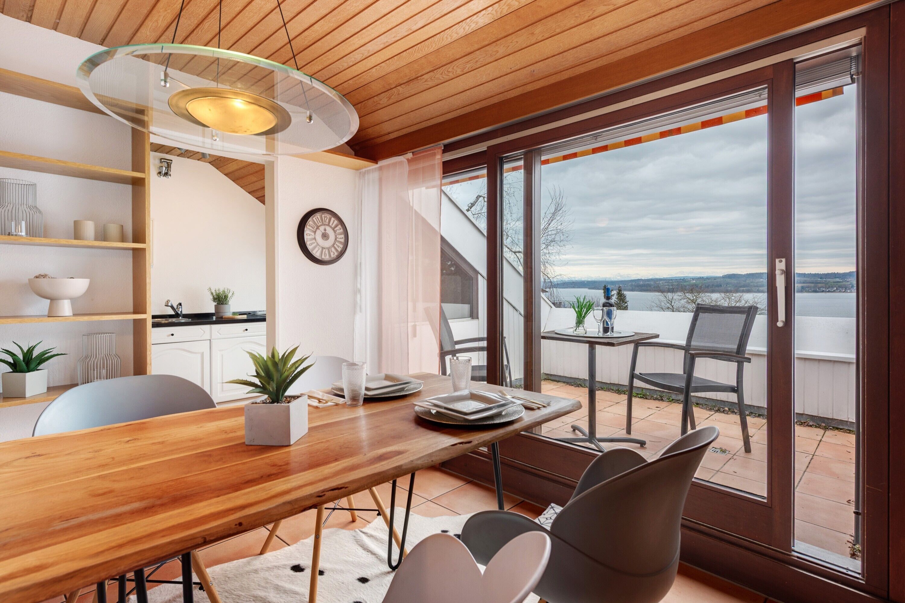 Un coin repas avec une table en bois, des chaises et une vue sur un lac, visible à travers de grandes portes-fenêtres coulissantes.