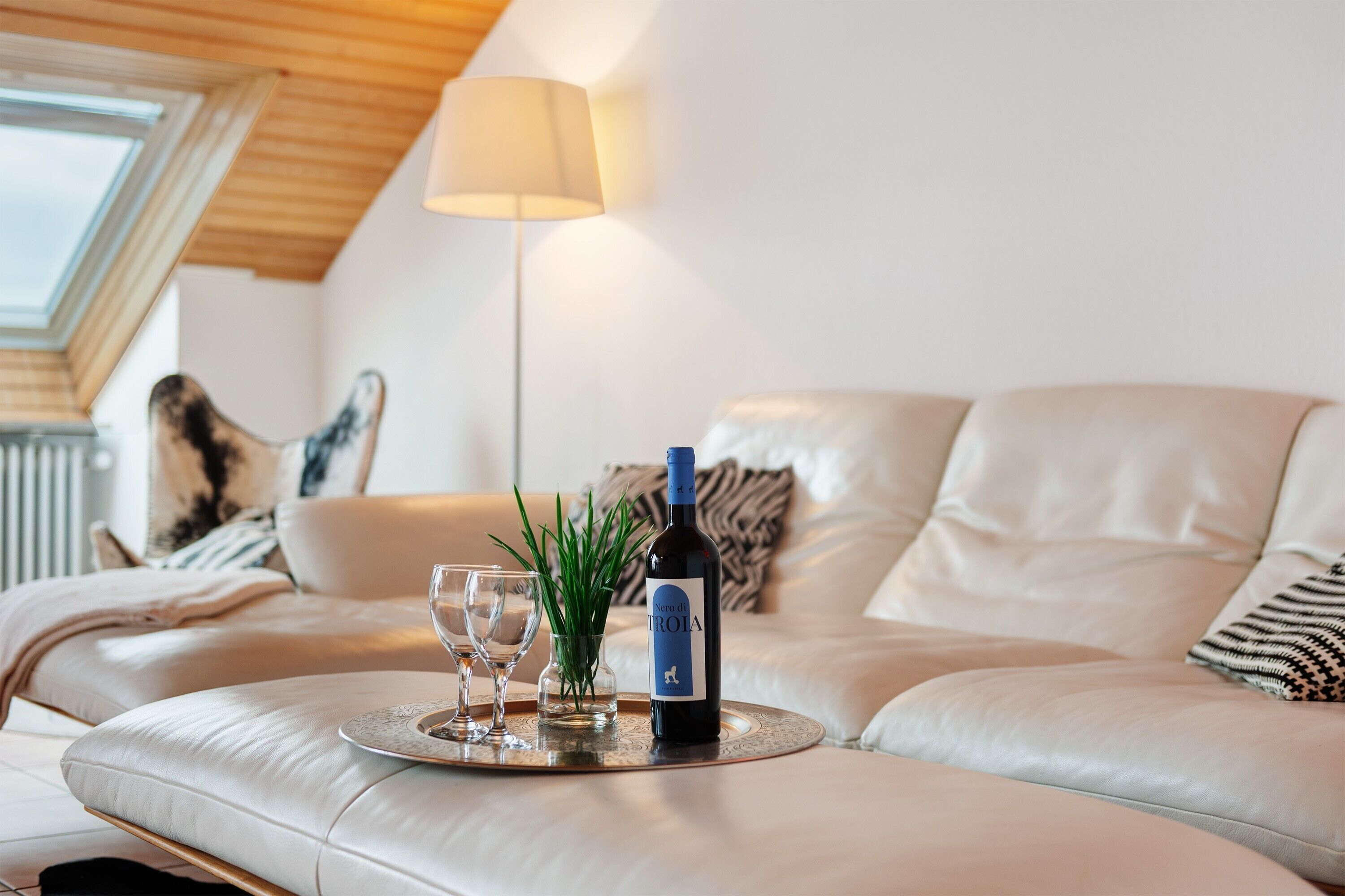 Ein modernes Wohnzimmer mit einer Couch, einem Glastisch mit Flasche und Gläsern, einer Lampe und einer Pflanze.