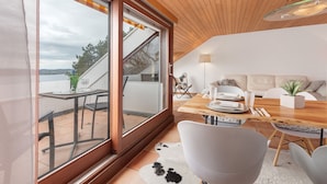 Un interior moderno con techo de madera, un comedor con mesa y sillas, un sofá, y vistas a través de amplios ventanales.