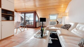Una sala de estar moderna con un sofá blanco, una mesa de centro y un televisor. Hay un comedor con sillas y una mesa, y una cocina con armarios y electrodomésticos.