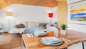Una sala moderna con techo de madera, una mesa de comedor con platos, vasos y cubiertos, un sofá con cojines, una lámpara de pie roja y un cuadro enmarcado en la pared.