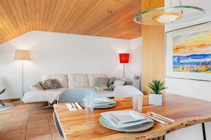 Una sala moderna con techo de madera, una mesa de comedor con platos, vasos y cubiertos, un sofá con cojines, una lámpara de pie roja y un cuadro enmarcado en la pared.