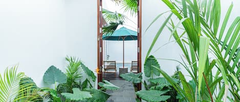 Un chemin de pierres mène à une porte en bois, surmontée d’un parapluie vert, le tout étant entouré d’une végétation tropicale luxuriante.