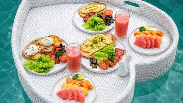 Una vajilla con platos de comida, un vaso de jugo y saleros y pimenteros sobre una bandeja de mimbre blanca junto a una piscina.
