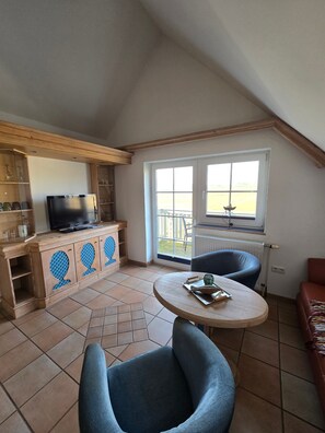 A room with a TV, wooden cabinets, a round table, a blue chair, and a view through a window. - Apartment 'Bachstelze' with Sea View, Shared Terrace and Wi-Fi (Mönchgut)
