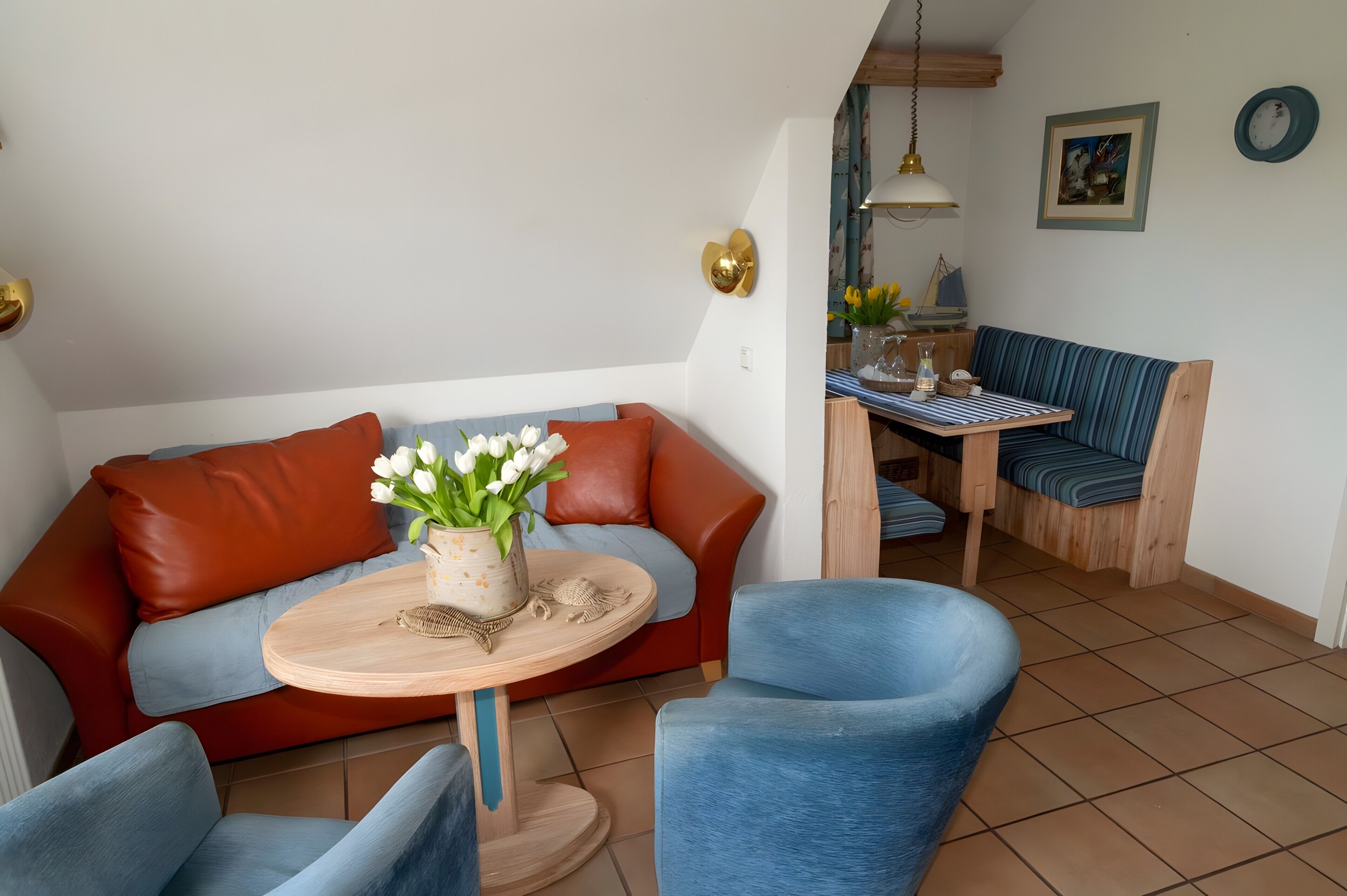 A living room with a sofa, a round table with a vase of flowers, a dining area with a striped bench, and a wall clock.