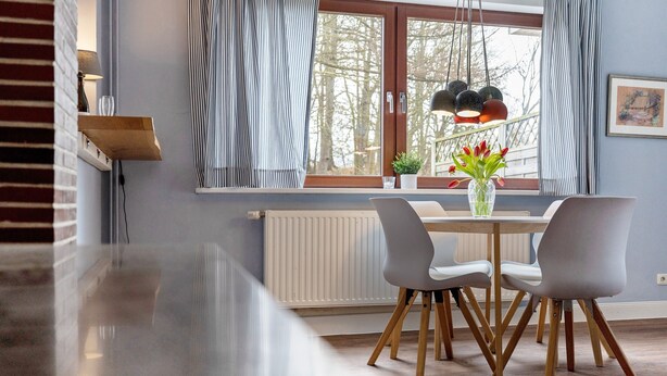 Ein Essbereich mit Tisch, Stühlen, einer Vase mit Blumen und einem Fenster mit Vorhängen.