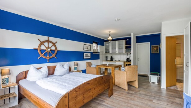 Un dormitorio con cama de madera, un escritorio y una silla, una kitchenette al fondo, y una decoración en tonos azul y blanco.