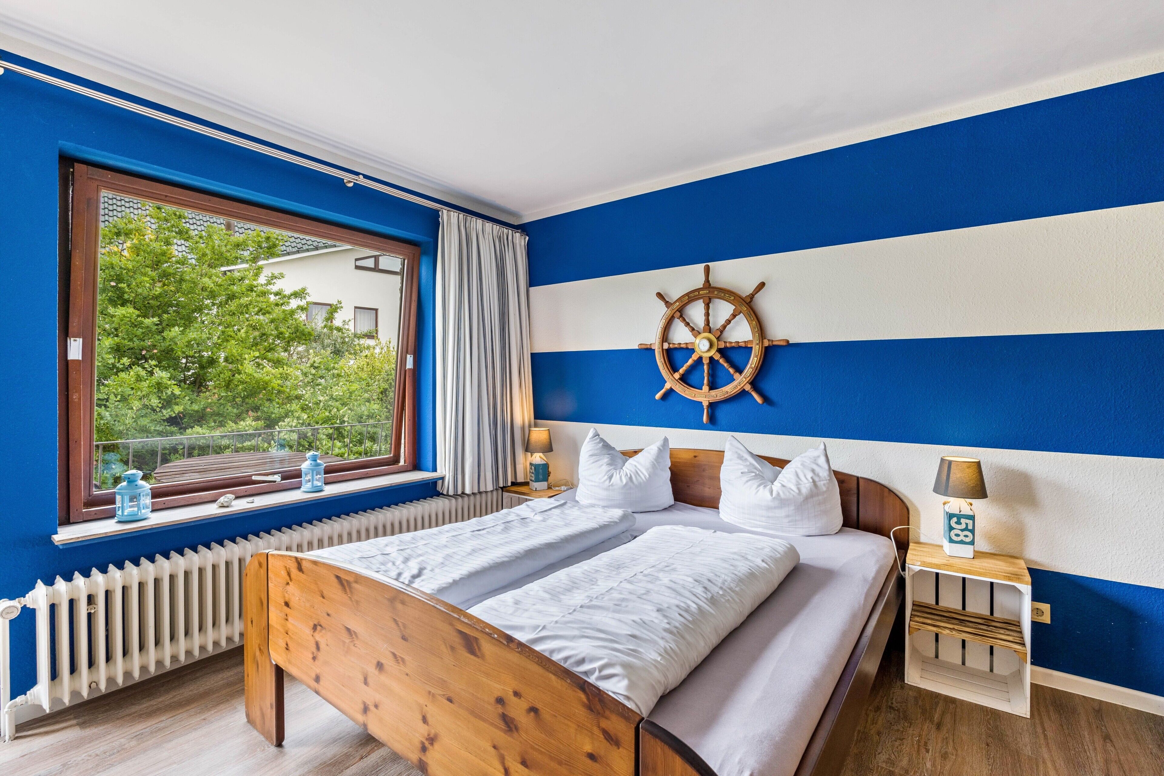 Quarto com uma grande janela, cama de madeira, decoração com um leme de navio e mesa de cabeceira com luminária.