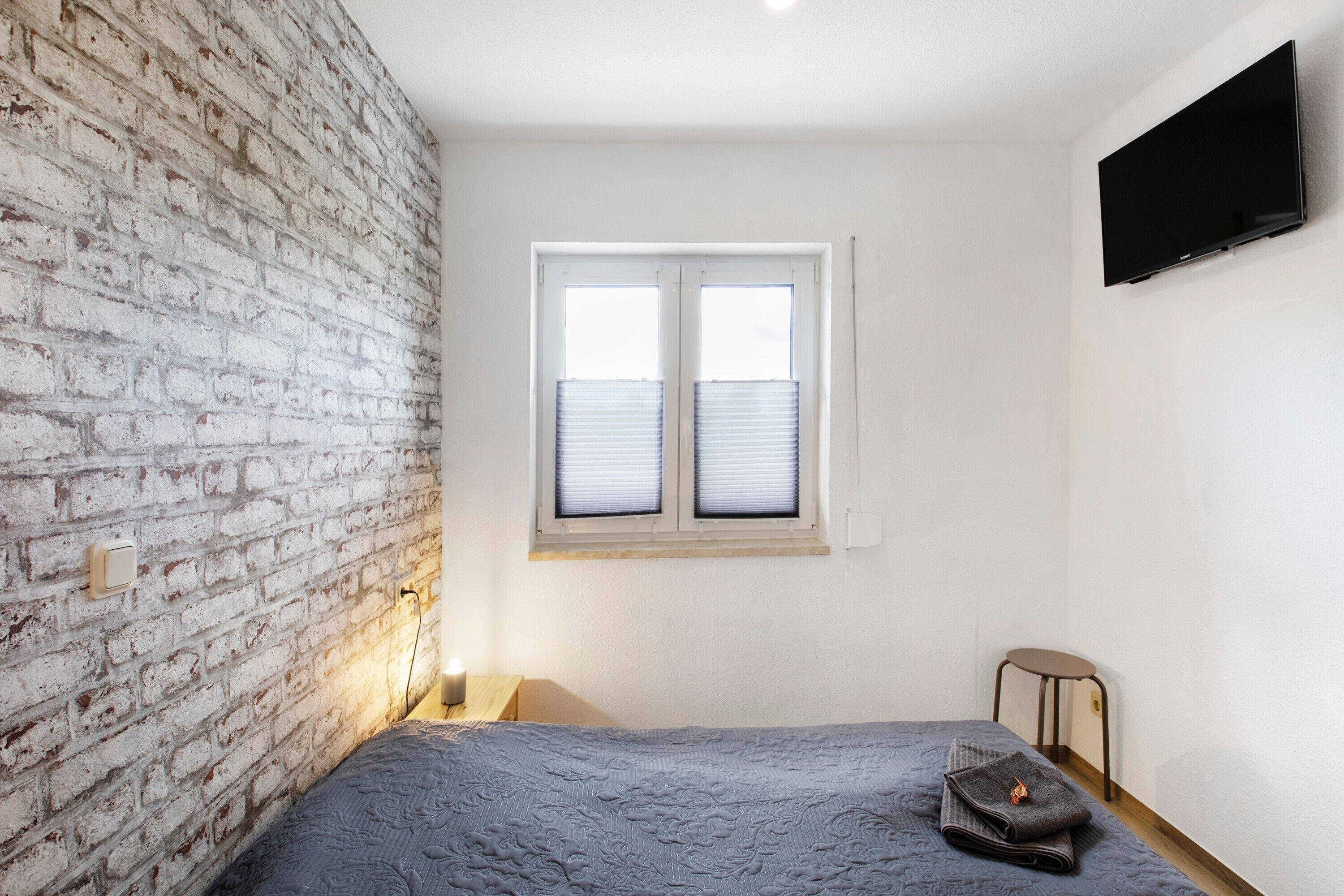 Une chambre avec un mur en briques, une fenêtre avec des stores, un lit avec une couverture, une table de chevet et un téléviseur fixé au mur.