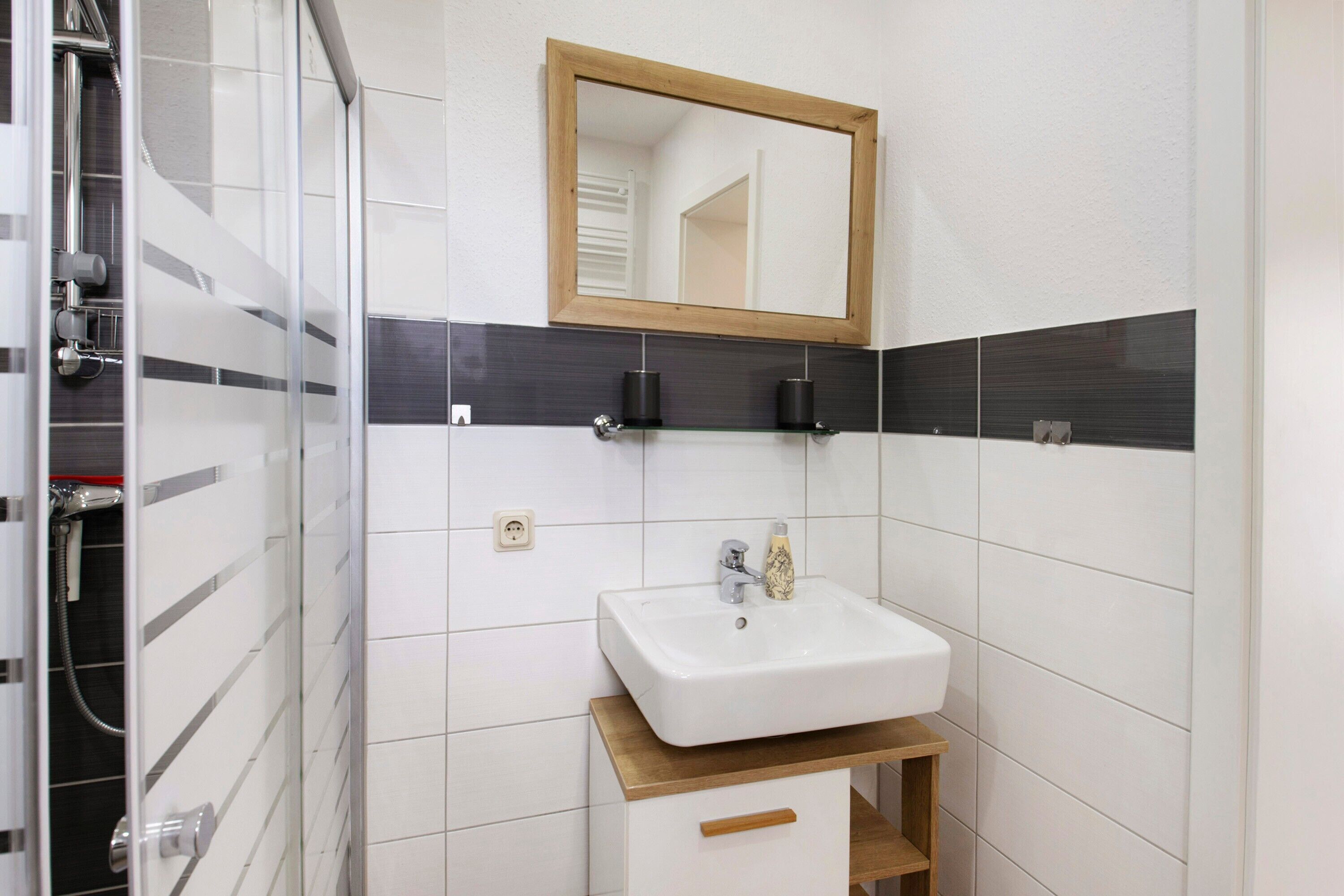 Ein Badezimmer mit weißem Waschbecken, einem Holzregal, einem Spiegel und einer Duschkabine.