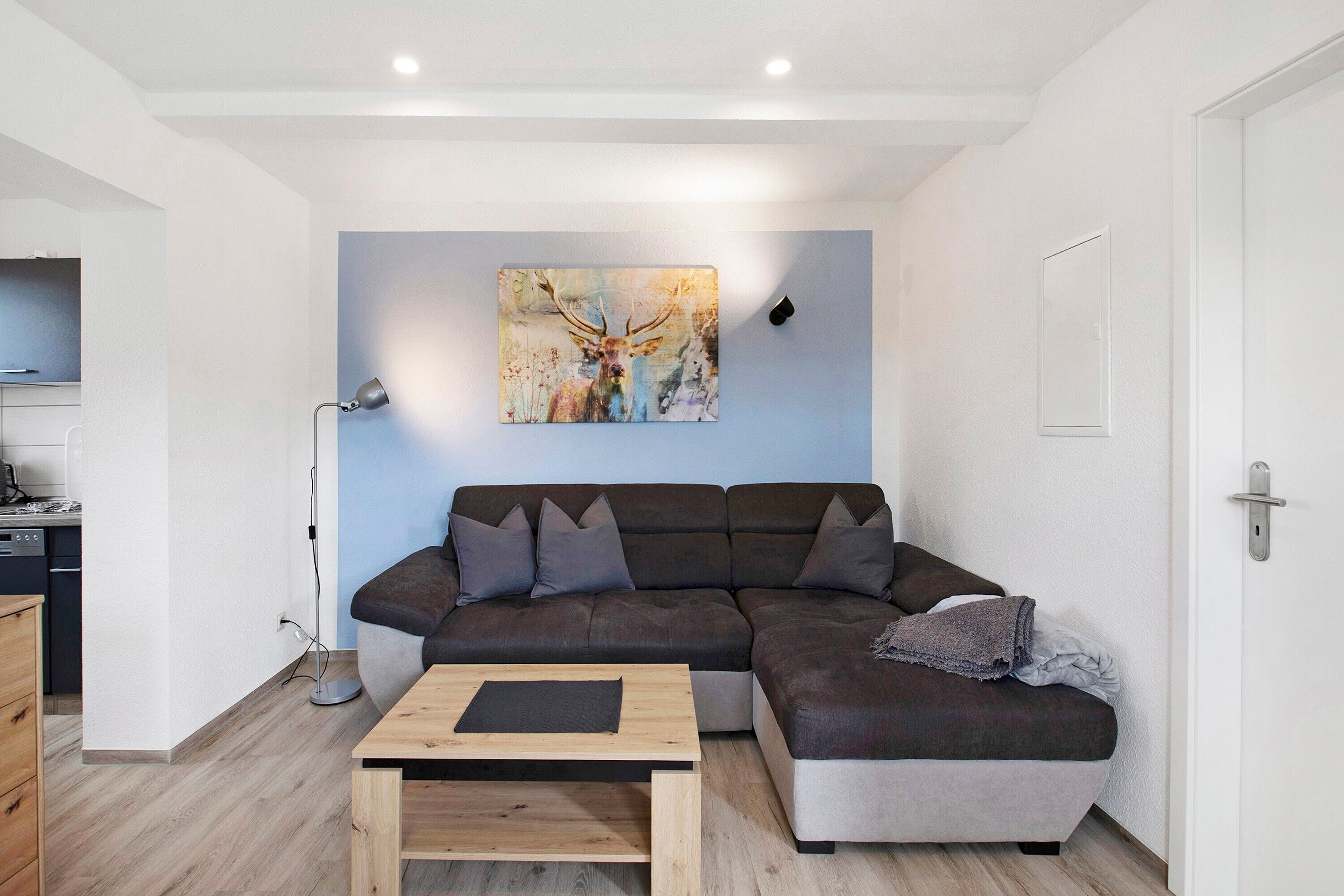 A modern living room with a sectional sofa, a coffee table, a painting of a deer, and a kitchen area visible to the left.