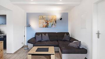 A modern living room with a sectional sofa, a coffee table, a painting of a deer, and a kitchen area visible to the left.