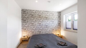 A bedroom with a brick wall, a bed with a dark cover, bedside tables with lamps, and a window with blinds.