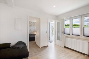 Un intérieur moderne avec un canapé, une porte blanche donnant sur un balcon et une vue sur la verdure à l’extérieur.