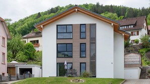 A two-story building with a flat roof, a stone wall in front, and a grassy area with a bench.