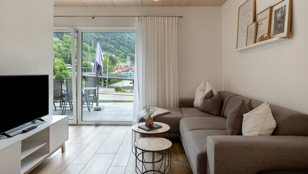 A modern living room with a flat-screen TV, a sofa, a small round table, and a balcony with a view of greenery and buildings.