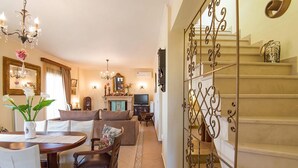 A spacious living room with a fireplace, a dining area with a round table, and a staircase with ornate railings. - Νikolaos Villa with Private Pool, Playground & BBQ (Rethymno)