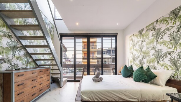 A bedroom with a bed, a wooden dresser, a staircase, and a large window with a view of buildings. - Lofty Comfort Stay-Relax by Soben Home (Phnom Penh)