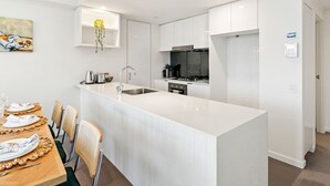 A modern kitchen with white cabinetry, a central island with a sink, and a dining area with a wooden table and chairs. - Unbeatable River & CBD Views Pool/2Car/Prime Spot (South Brisbane)