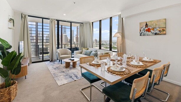 A modern living room with a dining area, large windows, and a city view. - Unbeatable River & CBD Views Pool/2Car/Prime Spot (South Brisbane)