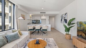 A modern living room with a sofa, coffee table, and dining area with chairs. There is a large window, a plant, and artwork on the walls. - Unbeatable River & CBD Views Pool/2Car/Prime Spot (South Brisbane)