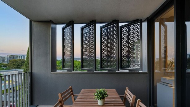 Een balkon met een houten tafel en stoelen, een plant in pot en uitzicht op groen en gebouwen.