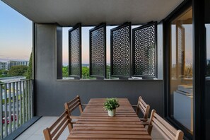 Een balkon met een houten tafel en stoelen, een plant in pot en uitzicht op groen en gebouwen.