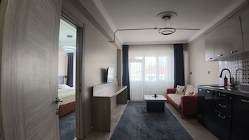 Una habitación de hotel moderna con cama, escritorio, sofá, zona de cocina y un ventanal con cortinas.