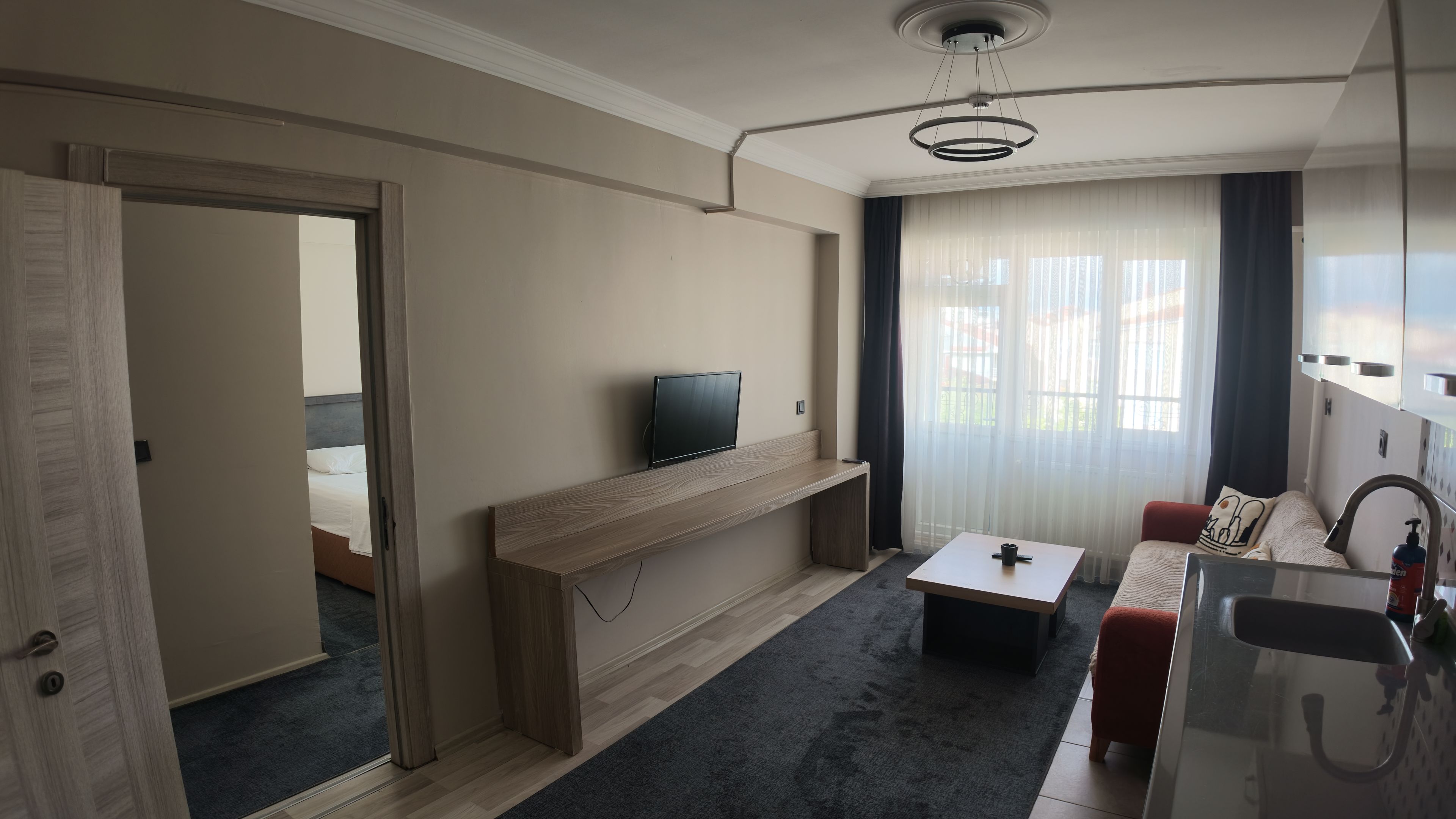 A modern hotel room with a television on a wooden console, a small table, a sofa, and a bathroom with a sink and mirror.
