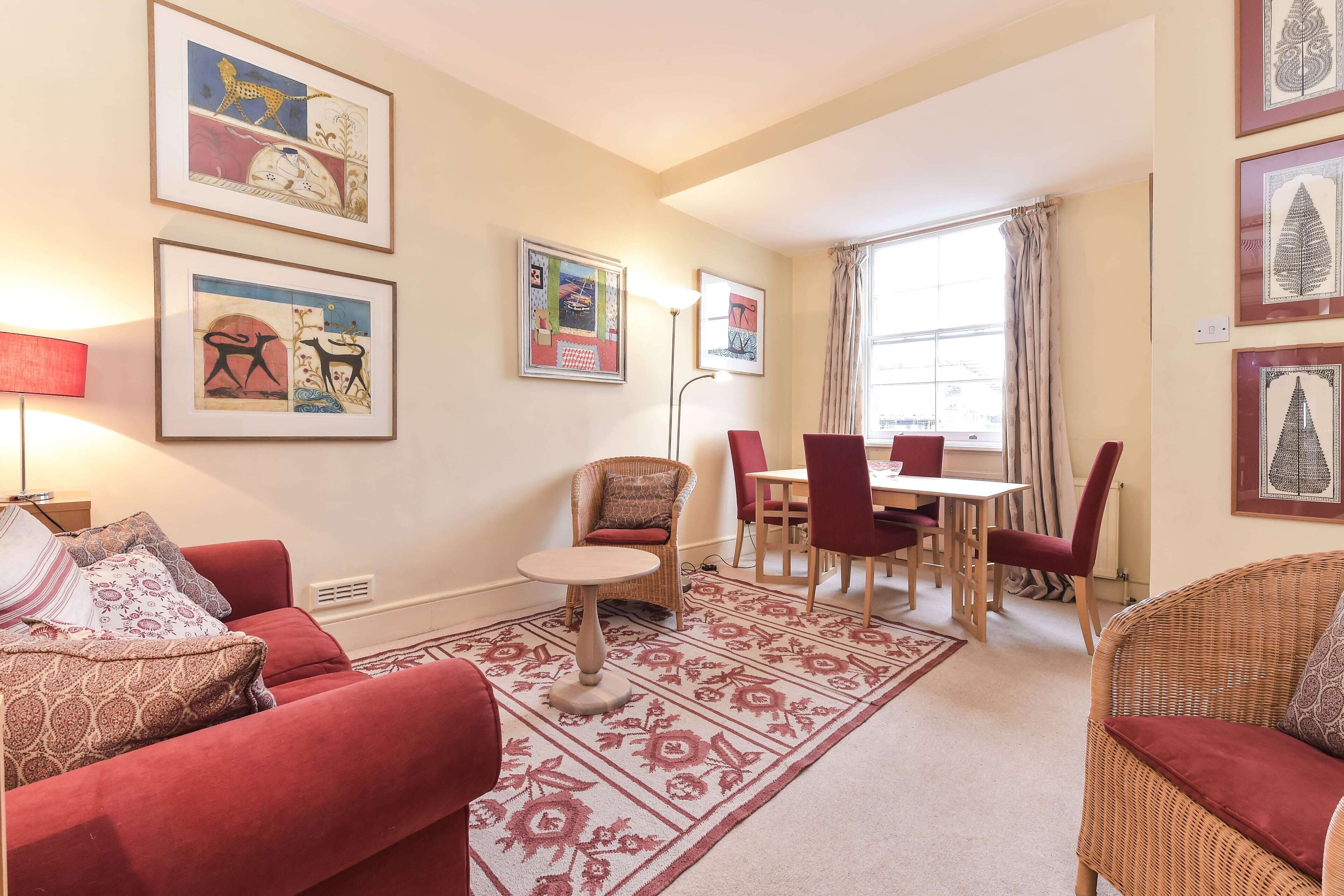 A living room with a red sofa, a round table, and a dining area with chairs. There are framed pictures on the walls and a window with curtains.