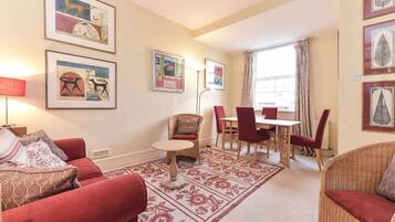 A living room with a red sofa, a round table, and a dining area with chairs. There are framed pictures on the walls and a window with curtains.
