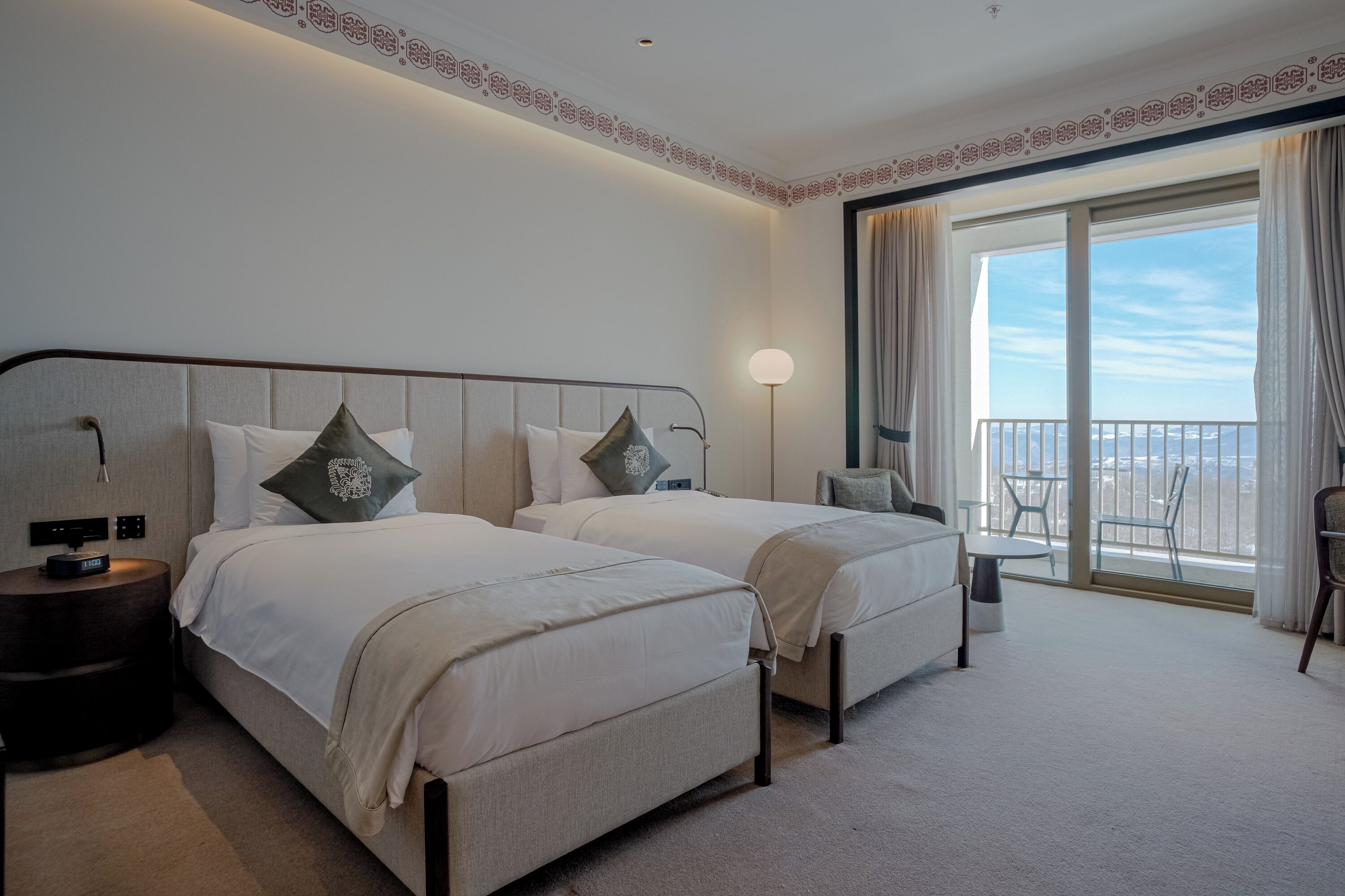 A hotel room with two beds, a bedside table, a lamp, a chair, and a balcony with a view.
