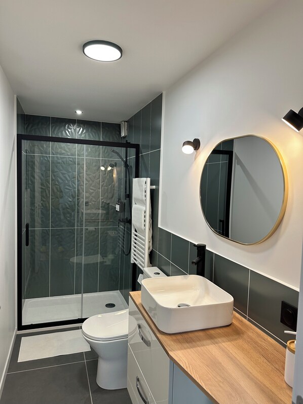 A modern bathroom with a glass shower enclosure, a white toilet, a vanity with a wooden countertop, and a round mirror with a gold frame.