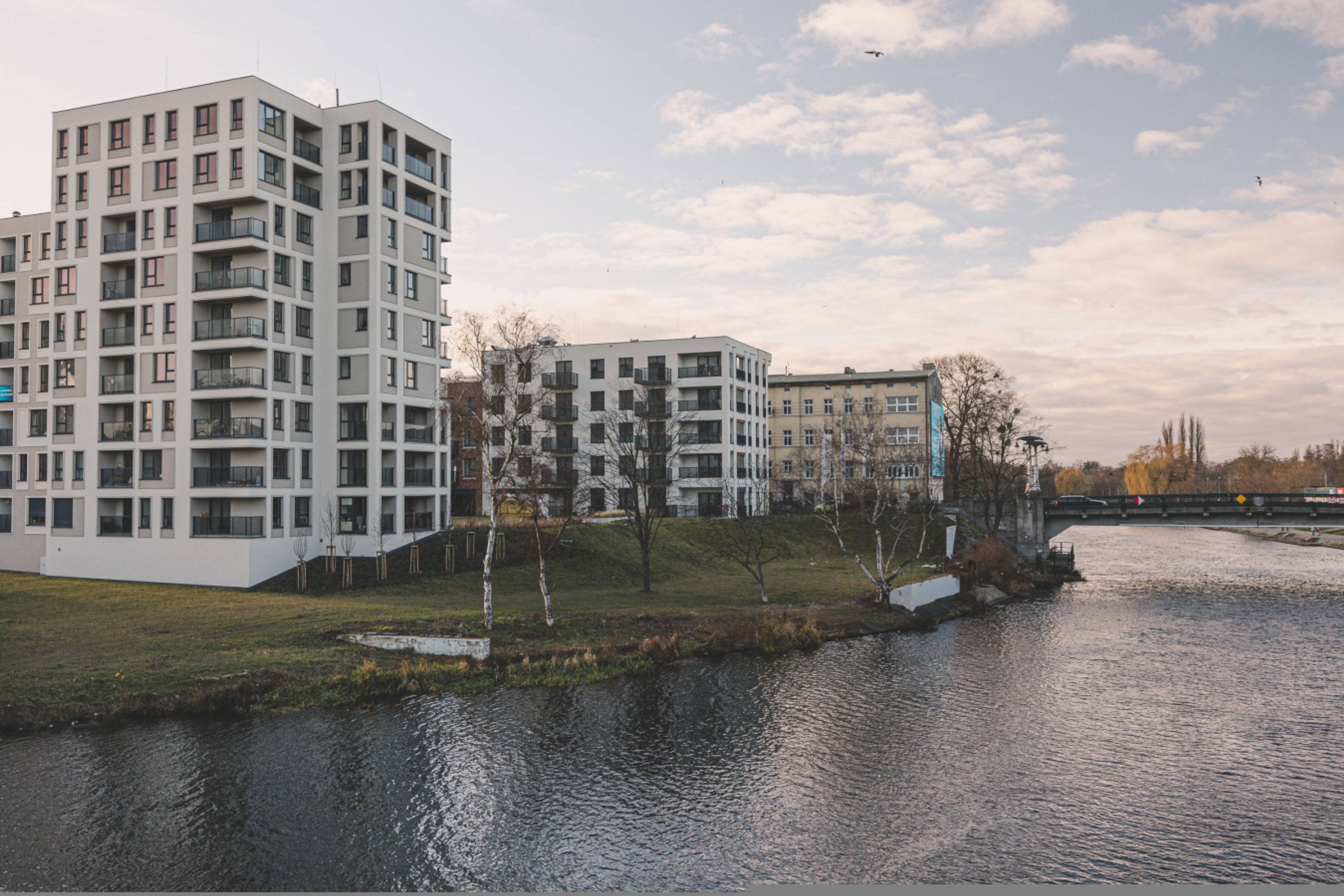 Een modern appartementencomplex met meerdere balkons, een rivier op de voorgrond en een brug op de achtergrond.