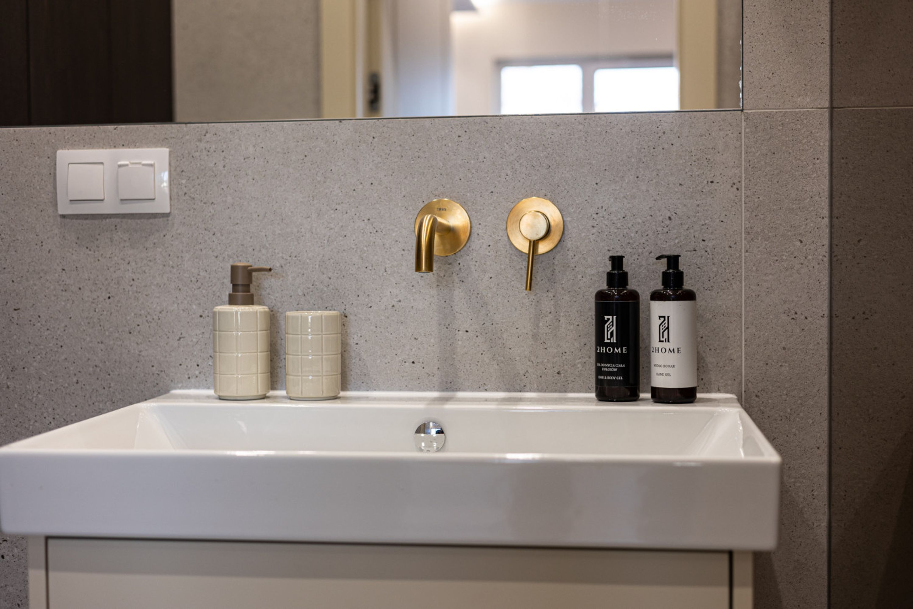 Un baño con un lavamanos blanco, dos grifos dorados y dos botellas de gel hidroalcohólico 2Home.
