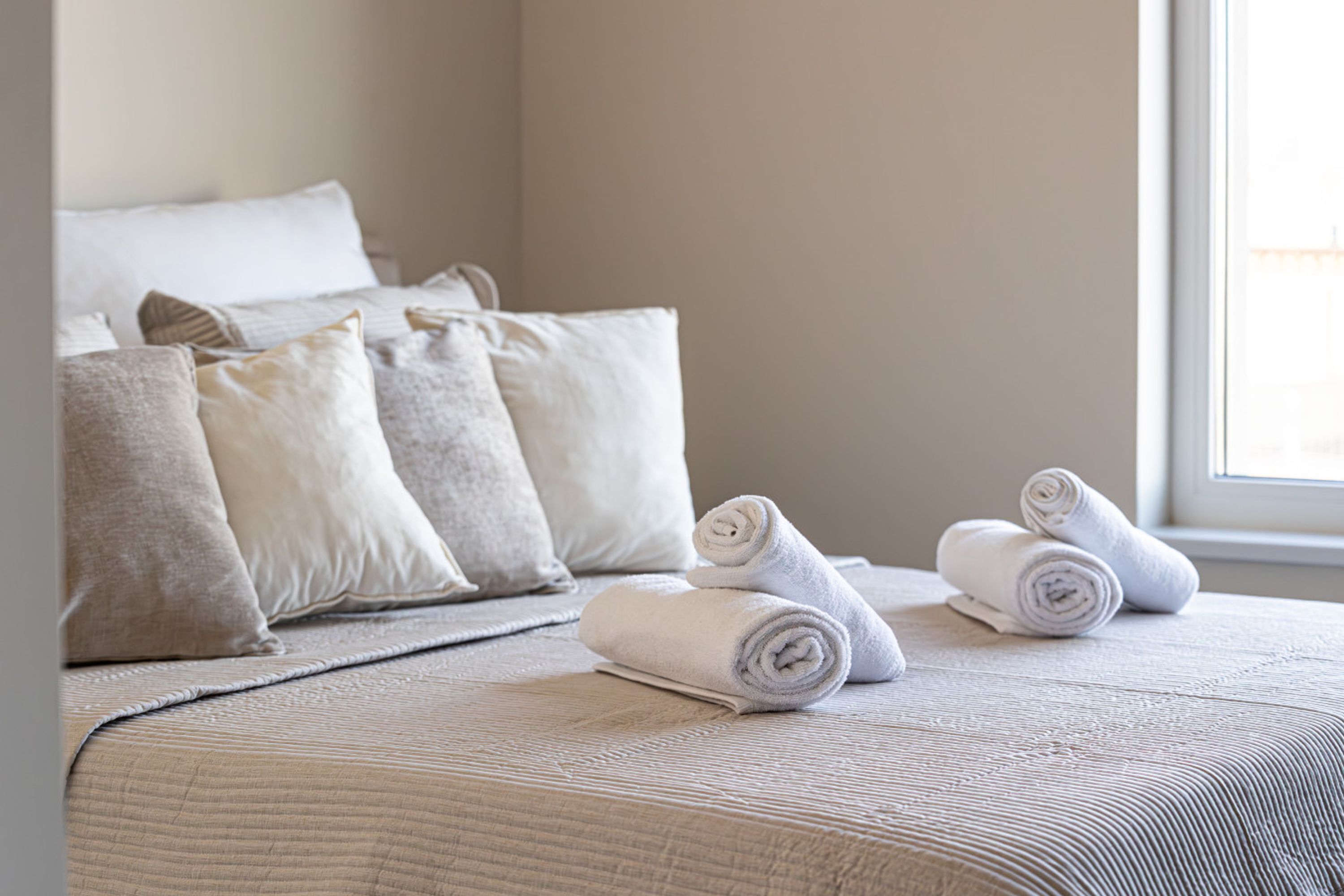 A neatly made bed with rolled towels and a view of the outside through a window.