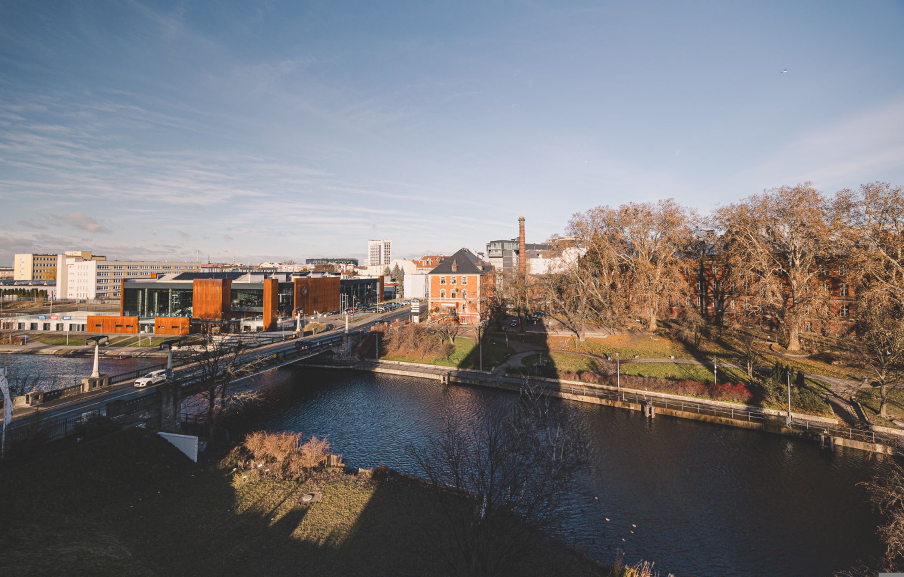 A riverside view of a cityscape with modern buildings, a bridge, and a park with leafless trees.