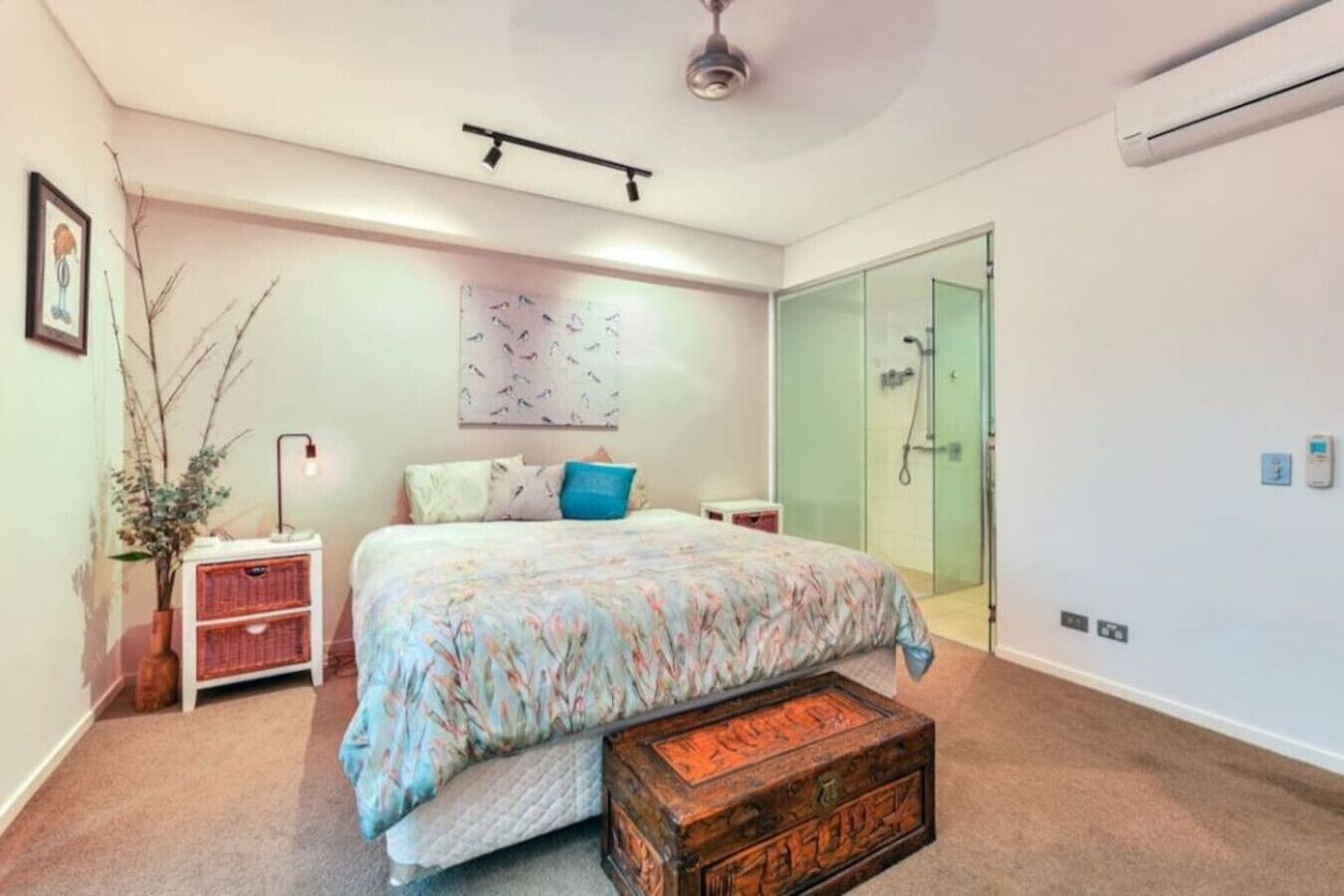 A bedroom with a bed, bedside tables, a dresser, a vase with branches, a wall art piece, a shower area, and an air conditioning unit.