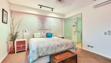 A bedroom with a bed, bedside tables, a dresser, a vase with branches, a wall art piece, a shower area, and an air conditioning unit.