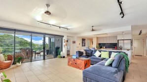 A spacious living area with a large blue sofa, a dining table, and a sliding glass door leading to a balcony with outdoor furniture. - Benji Zen Retreat Unique Wavepool Adults Only (Darwin)